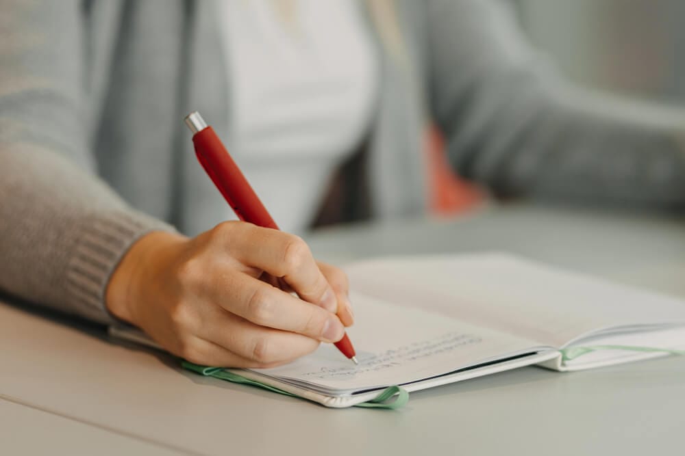 A woman writing in a journal with a red pen.