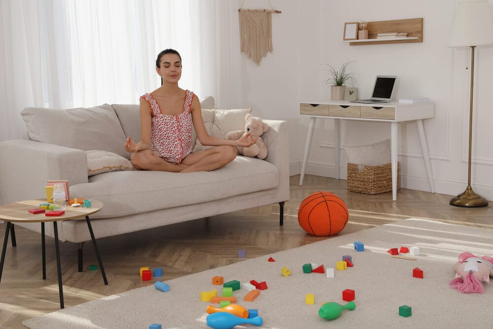 A woman sits in the lotus position on a sofa in a room with toys all over the floor.