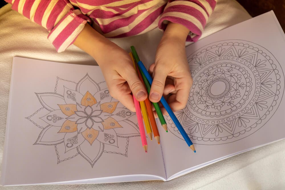 A woman holds different coloring pencils over a mandala coloring book.