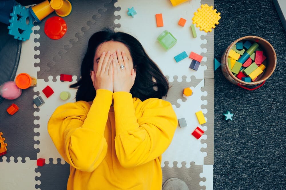A stressed mom lies on her back on a toy mat, with her hands covering her face.