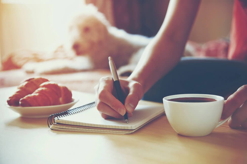 A woman sits at a table, writing in a notebook, about to drink a cup of coffee.