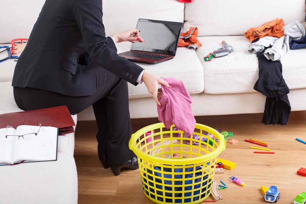 A woman is putting laundry in a basket while working on a laptop. Children's toys are strewn on the floor.