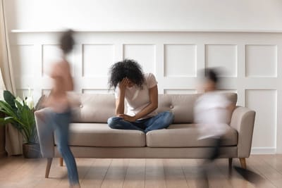 An overstimulated mom sits in the middle of the sofa with her head in her hands while two kids run around her.
