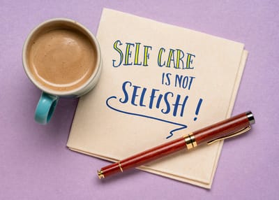 A cup of coffee and a red pen are next to a napkin with the message "Self Care is Not Selfish!"