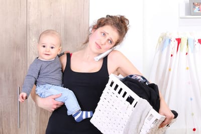 A woman is holding her child in one arm, a basket of laundry in the other, and has a phone tucked under her ear.